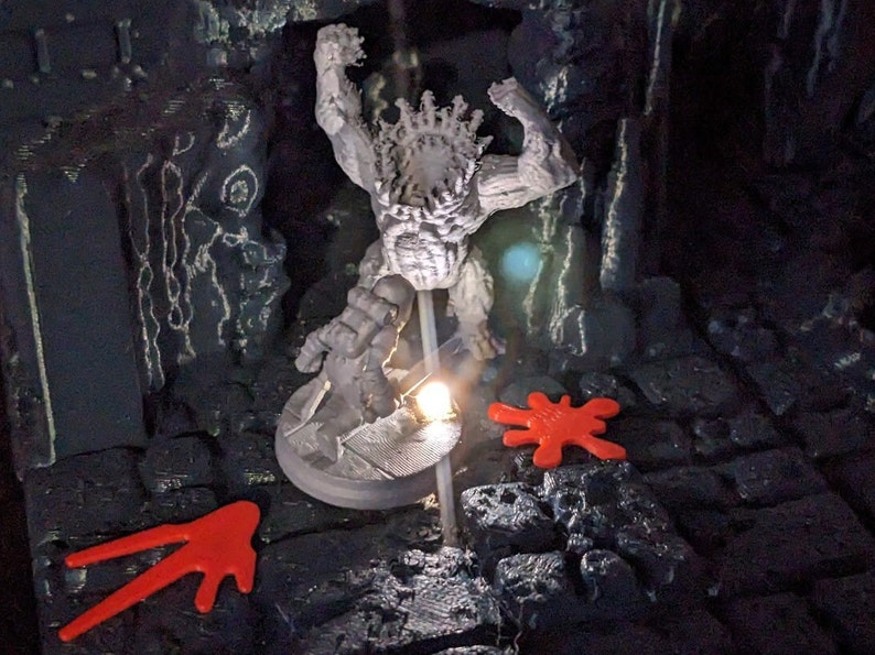 May include: A gray figurine of a muscular figure with a crown, posed in a dramatic stance. The figure is on a round base, illuminated by a small light. Two red, abstract shapes are on the dark, textured surface.