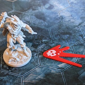 May include: A gray miniature figure of a warrior with a spiked club and a knife, standing on a round base. A red arrow-shaped game piece with a white skull icon is next to the figure. The game board has a hexagonal tile pattern.