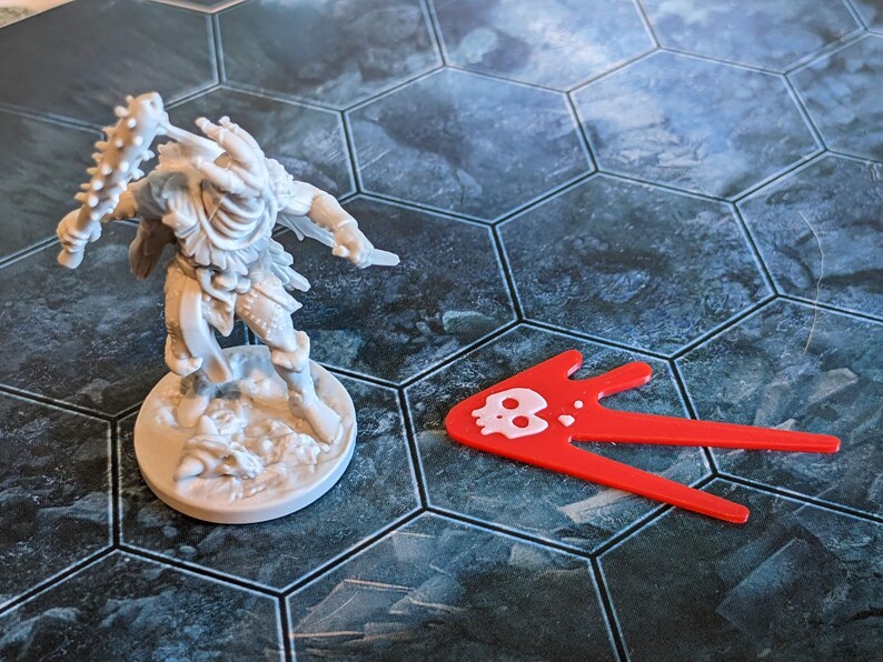 May include: A gray miniature figure of a warrior with a spiked club and a knife, standing on a round base. A red arrow-shaped game piece with a white skull icon is next to the figure. The game board has a hexagonal tile pattern.