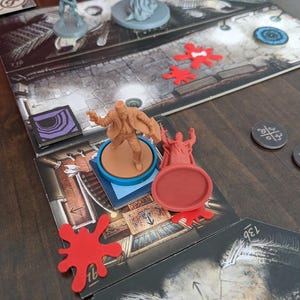 May include: A board game setup featuring game pieces and a game board. The game board has a detailed illustration of a room. Several game pieces are visible, including miniature figures and red tokens. The game is set on a dark wooden surface.