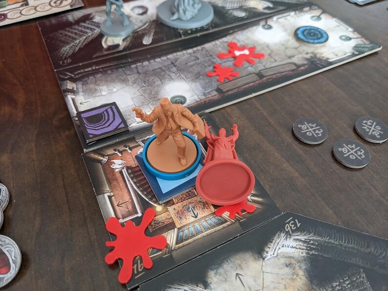 May include: A board game setup featuring game pieces and a game board. The game board has a detailed illustration of a room. Several game pieces are visible, including miniature figures and red tokens. The game is set on a dark wooden surface.