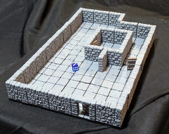 May include: A white painted miniature dungeon tile with a stone wall border and a small room with a door. A blue die is on the floor of the tile.
