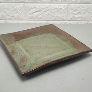 May include: Square ceramic plate with a brown rim and a green center. The plate has a rustic, handmade appearance. The plate is suitable for serving appetizers or small dishes.