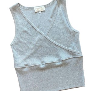 May include: A sleeveless, silver-gray wrap-style top with a V-neckline. The top has a ribbed texture and a fitted silhouette. The label on the top reads "cache" and "L".