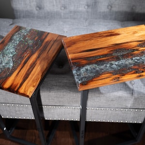 May include: Two wooden side tables with a river-like design. The tables have a dark wood grain with a blue-green epoxy resin center. The tables have black metal legs and are placed on a gray upholstered sofa.