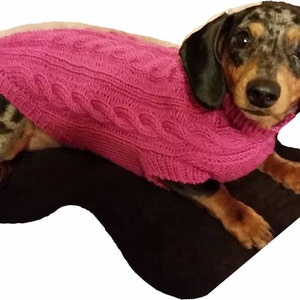 Puede incluir: Un pequeño dachshund de pelo corto con un suéter de punto rosa de cable. El perro está acostado sobre una superficie negra y mirando directamente a la cámara.