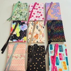 May include: Nine rectangular fabric pouches arranged in a grid, each featuring a unique pattern including succulents, cherries, floral prints, abstract paint splatters, ikat, paw prints, stars, and retro boomboxes, all with attached ribbon ties.