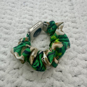 May include: A green and tan sloth-patterned scrunchie with an Apple Watch attached. The scrunchie is made of fabric and has a gathered design. The watch is silver and has a digital display.