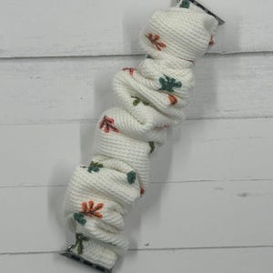 May include: White Apple Watch band with a textured knit design. The band features a repeating pattern of orange and green leaves. The band has silver connectors on each end.