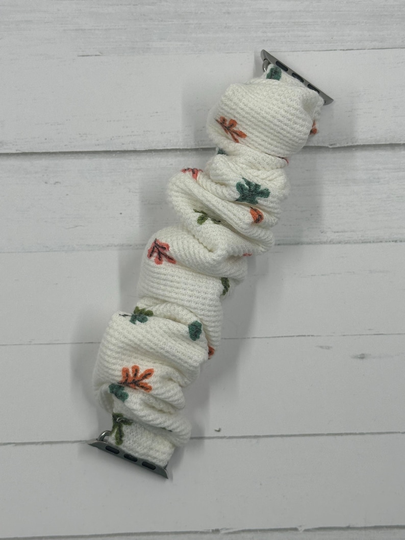 May include: White Apple Watch band with a textured knit design. The band features a repeating pattern of orange and green leaves. The band has silver connectors on each end.