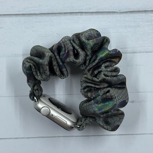 May include: A silver smartwatch with a dark gray scrunchie band. The scrunchie band has a textured, iridescent pattern with hints of green, blue, and purple. The watch is attached to the band with silver connectors.