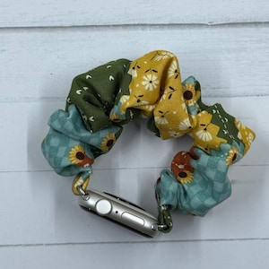 May include: A colorful Apple Watch band featuring a patchwork design with green, yellow, and teal fabrics. The band is adorned with floral patterns, including sunflowers and small white flowers. The watch band is attached to a silver Apple Watch.