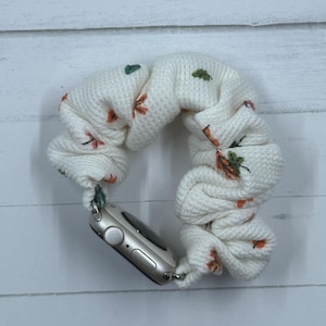 May include: Cream-colored Apple Watch scrunchie band with a waffle knit texture. The band is adorned with small orange and green floral embroidery. The watch face is silver.