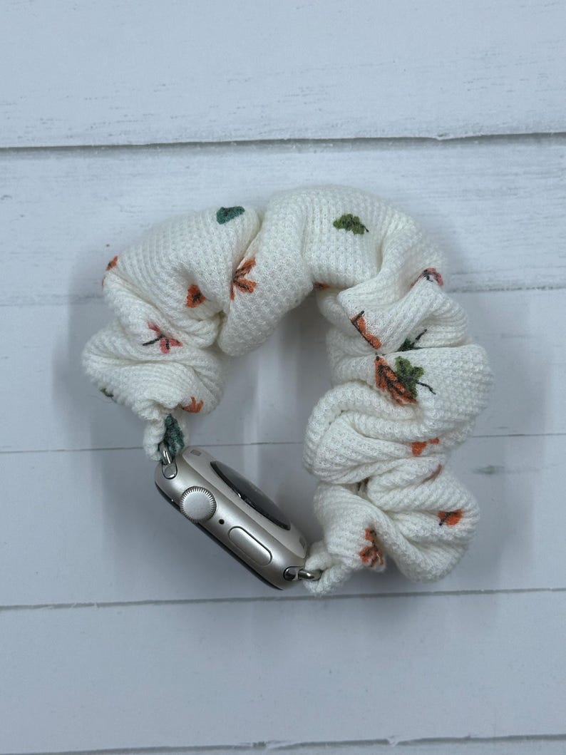 May include: Cream-colored Apple Watch scrunchie band with a waffle knit texture. The band is adorned with small orange and green floral embroidery. The watch face is silver.