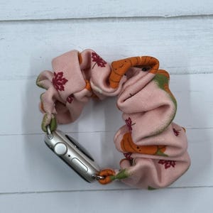 May include: A pink scrunchie with an orange pumpkin and burgundy leaf pattern, attached to a silver smartwatch. The scrunchie has green trim and is designed to hold the watch securely. The watch band is a stylish accessory.