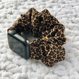 May include: A black smartwatch with a leopard print scrunchie band. The scrunchie is brown with black spots. The watch is on a white, textured surface.