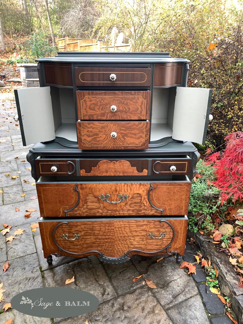 Puede incluir: Una c&oacute;moda vintage con un acabado de pintura verde oscuro y cajones de chapa de madera. La c&oacute;moda tiene una secci&oacute;n superior con dos puertas que se abren para revelar tres cajones. La secci&oacute;n inferior tiene cuatro cajones con herrajes ornamentados. La c&oacute;moda est&aacute; sentada en un patio de piedra.