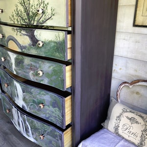 May include: A dark brown dresser with five drawers. Each drawer is painted with a different nature scene, including a waterfall, trees, and a bridge. The dresser is in a room with a white-painted wall and a white bench with a pillow that says "Paris".