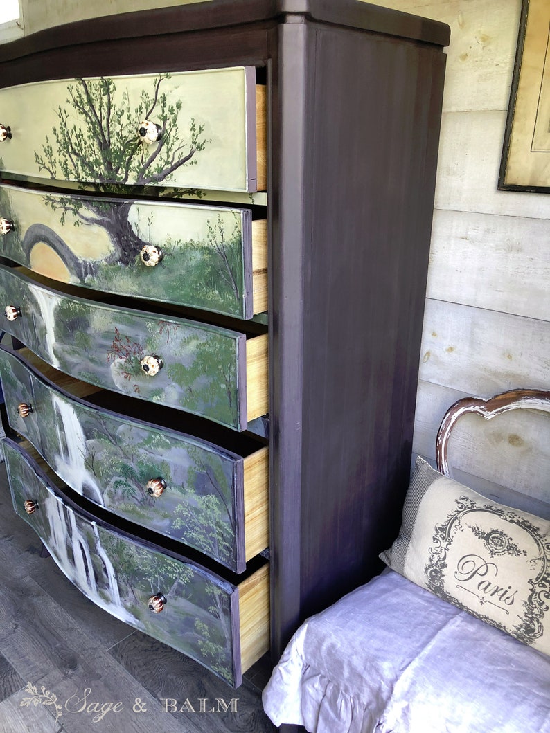 May include: A dark brown dresser with five drawers. Each drawer is painted with a different nature scene, including a waterfall, trees, and a bridge. The dresser is in a room with a white-painted wall and a white bench with a pillow that says "Paris".