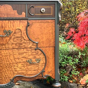 Puede incluir: Una c&oacute;moda de madera vintage con tallas intrincadas y un acabado de pintura verde oscuro. La c&oacute;moda tiene varios cajones con herrajes de lat&oacute;n ornamentados y un patr&oacute;n de veta de madera. La c&oacute;moda est&aacute; fotografiada al aire libre con follaje oto&ntilde;al en el fondo.
