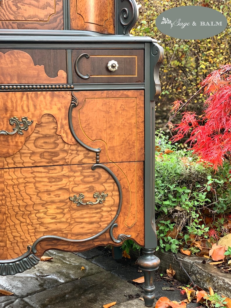 Puede incluir: Una c&oacute;moda de madera vintage con tallas intrincadas y un acabado de pintura verde oscuro. La c&oacute;moda tiene varios cajones con herrajes de lat&oacute;n ornamentados y un patr&oacute;n de veta de madera. La c&oacute;moda est&aacute; fotografiada al aire libre con follaje oto&ntilde;al en el fondo.