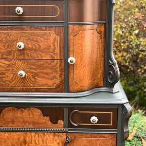 Puede incluir: Una c&oacute;moda vintage con un acabado de pintura gris oscuro y detalles de veta de madera. La c&oacute;moda tiene cuatro cajones y una puerta de armario con un dise&ntilde;o curvo. La c&oacute;moda tiene herrajes decorativos de lat&oacute;n y pomos de porcelana blanca.