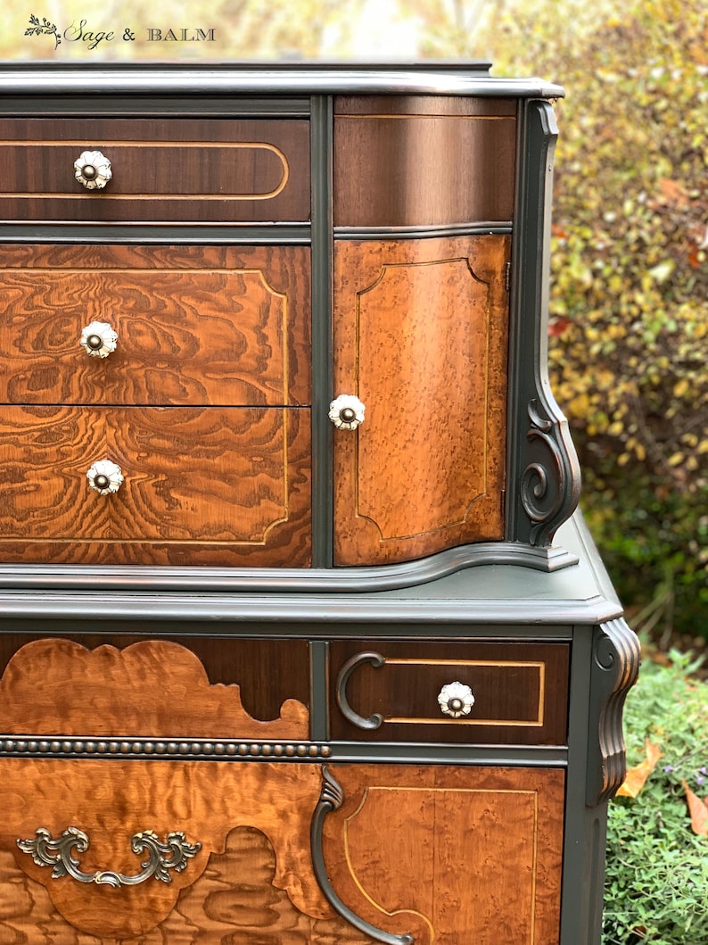 Puede incluir: Una c&oacute;moda vintage con un acabado de pintura gris oscuro y detalles de veta de madera. La c&oacute;moda tiene cuatro cajones y una puerta de armario con un dise&ntilde;o curvo. La c&oacute;moda tiene herrajes decorativos de lat&oacute;n y pomos de porcelana blanca.