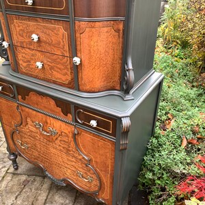 Puede incluir: Una c&oacute;moda vintage con un acabado de pintura verde oscuro y cajones de chapa de madera. La c&oacute;moda tiene detalles ornamentados y herrajes decorativos. La c&oacute;moda est&aacute; sentada en un patio de piedra.