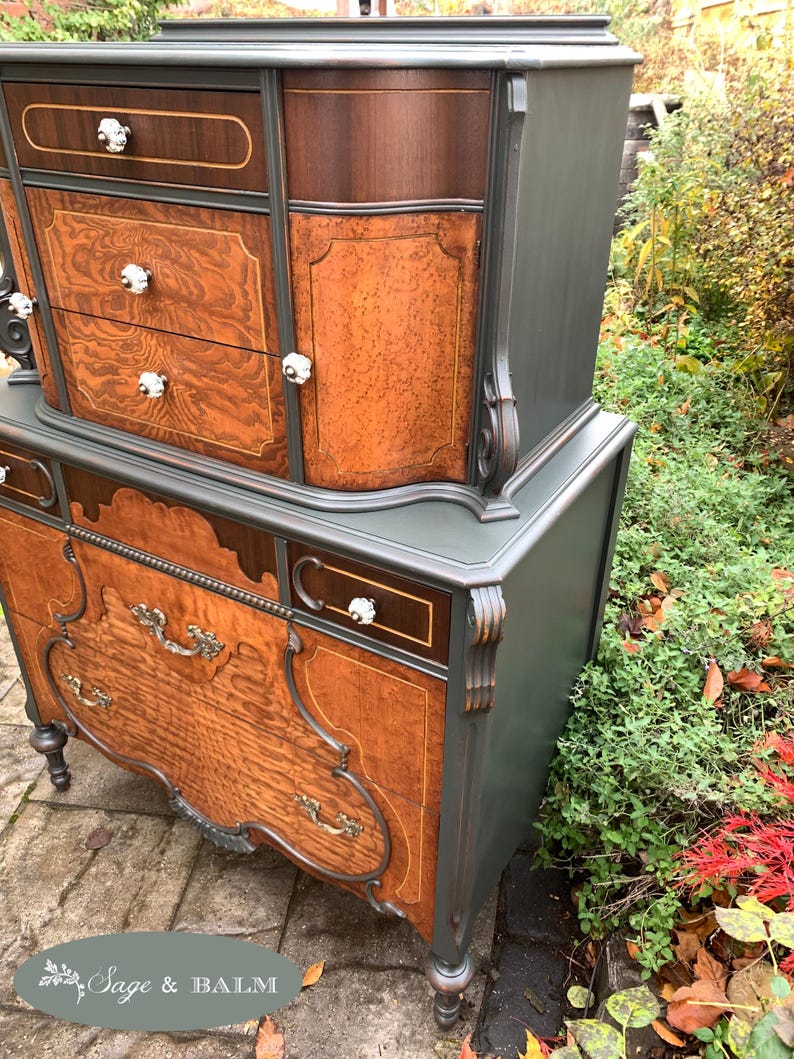 Puede incluir: Una c&oacute;moda vintage con un acabado de pintura verde oscuro y cajones de chapa de madera. La c&oacute;moda tiene detalles ornamentados y herrajes decorativos. La c&oacute;moda est&aacute; sentada en un patio de piedra.