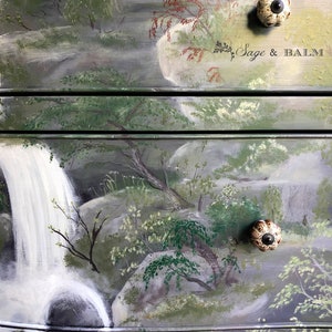 May include: A hand-painted dresser with a waterfall and forest scene. The dresser has two drawers with ceramic knobs. The top drawer has the text "Sage & Balm" painted on it.
