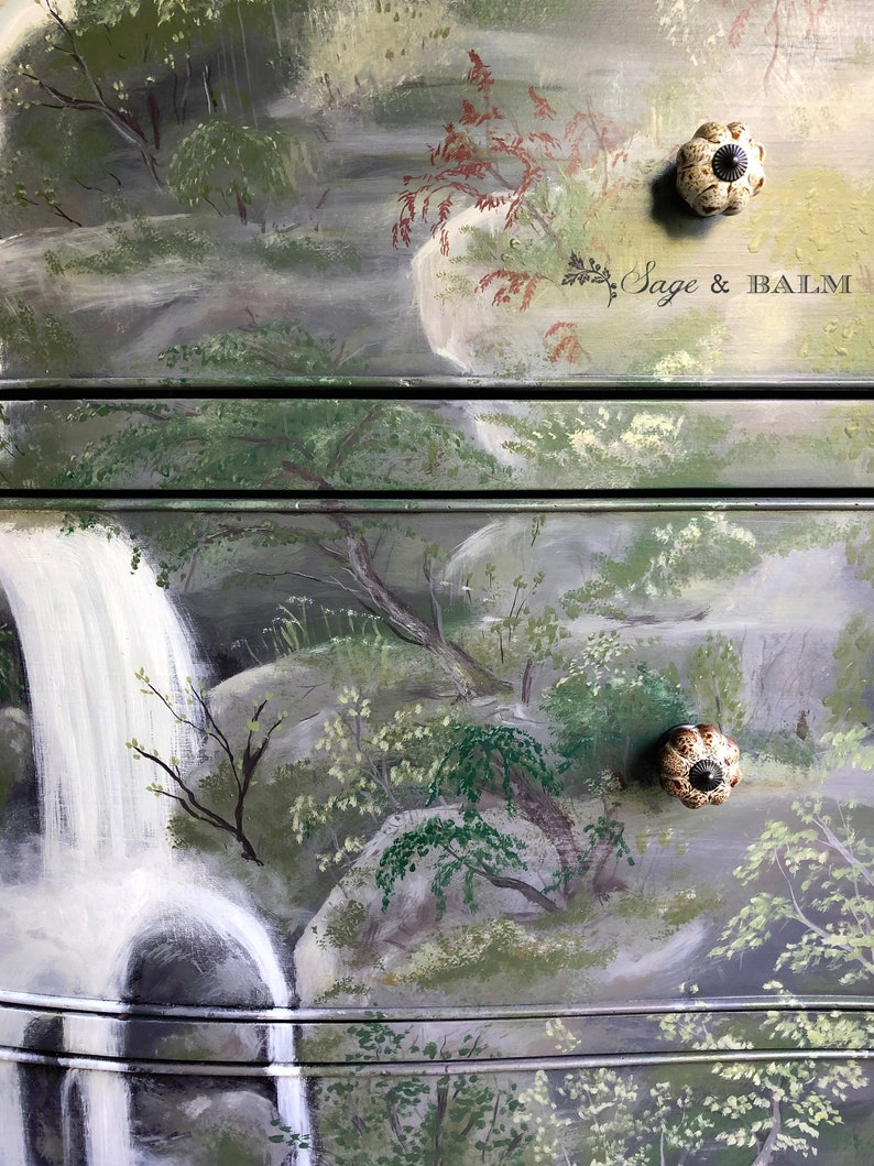 May include: A hand-painted dresser with a waterfall and forest scene. The dresser has two drawers with ceramic knobs. The top drawer has the text "Sage & Balm" painted on it.
