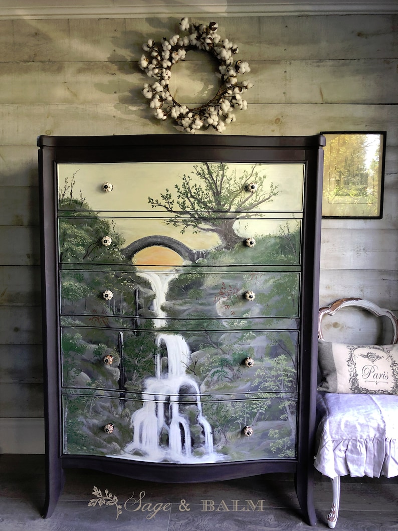 May include: A dark brown dresser with a hand-painted scene of a waterfall and a bridge. The drawers have white ceramic knobs. The dresser is in front of a white wood wall with a cotton wreath hanging above it.