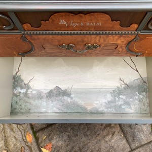 Puede incluir: Una c&oacute;moda de madera con un caj&oacute;n interior pintado que representa un paisaje mar&iacute;timo. El caj&oacute;n est&aacute; abierto y el interior est&aacute; pintado con un cielo azul claro, nubes blancas y una costa verde y marr&oacute;n. La c&oacute;moda tiene un acabado de madera oscura y herrajes ornamentados. El texto "Sage & Balm" est&aacute; grabado en la parte superior de la c&oacute;moda.