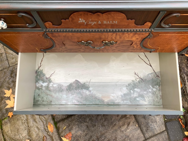 Puede incluir: Una c&oacute;moda de madera con un caj&oacute;n interior pintado que representa un paisaje mar&iacute;timo. El caj&oacute;n est&aacute; abierto y el interior est&aacute; pintado con un cielo azul claro, nubes blancas y una costa verde y marr&oacute;n. La c&oacute;moda tiene un acabado de madera oscura y herrajes ornamentados. El texto "Sage & Balm" est&aacute; grabado en la parte superior de la c&oacute;moda.