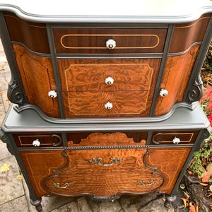 Puede incluir: Una c&oacute;moda vintage con un acabado de pintura gris oscuro y detalles de veta de madera. La c&oacute;moda tiene una parte superior curva y herrajes ornamentados. La c&oacute;moda est&aacute; hecha de madera y tiene un aspecto desgastado.