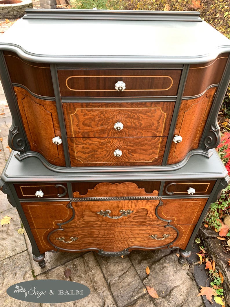 Puede incluir: Una c&oacute;moda vintage con un acabado de pintura gris oscuro y detalles de veta de madera. La c&oacute;moda tiene una parte superior curva y herrajes ornamentados. La c&oacute;moda est&aacute; hecha de madera y tiene un aspecto desgastado.