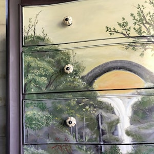 May include: A dark brown dresser with five drawers, each painted with a different landscape scene. The top drawer features a light yellow background with a green tree and a white ceramic knob. The second drawer has a green and brown landscape with a stone bridge and a white ceramic knob. The third drawer features a waterfall scene with a white ceramic knob. The fourth drawer has a green and brown landscape with a white ceramic knob. The bottom drawer has a green and brown landscape with a white ceramic knob.