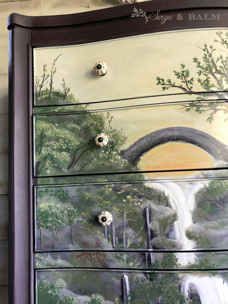 May include: A dark brown dresser with five drawers, each painted with a different landscape scene. The top drawer features a light yellow background with a green tree and a white ceramic knob. The second drawer has a green and brown landscape with a stone bridge and a white ceramic knob. The third drawer features a waterfall scene with a white ceramic knob. The fourth drawer has a green and brown landscape with a white ceramic knob. The bottom drawer has a green and brown landscape with a white ceramic knob.