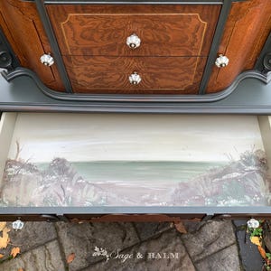 Puede incluir: Una c&oacute;moda vintage con un acabado gris oscuro y detalles ornamentados. El caj&oacute;n inferior est&aacute; abierto, revelando una escena pintada a mano de una playa con un mar verde y acantilados grises.