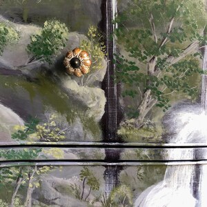 May include: A hand-painted dresser with a waterfall scene. The dresser has two drawers with brown ceramic knobs. The top drawer has the text "Sage & Balm" painted on it.