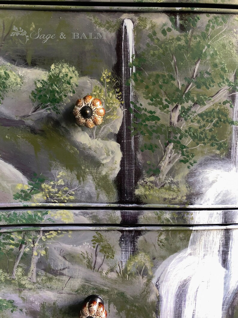 May include: A hand-painted dresser with a waterfall scene. The dresser has two drawers with brown ceramic knobs. The top drawer has the text "Sage & Balm" painted on it.