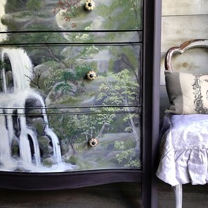 May include: A dark purple dresser with four drawers painted with a scenic waterfall landscape. The drawers have decorative knobs with a black center and a cream-colored rim. The dresser is sitting on a wood floor.