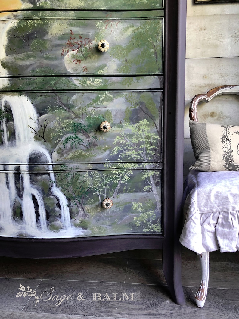 May include: A dark purple dresser with four drawers painted with a scenic waterfall landscape. The drawers have decorative knobs with a black center and a cream-colored rim. The dresser is sitting on a wood floor.