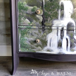 May include: A dark purple dresser with a hand-painted waterfall scene on the drawers. The dresser has a curved bottom and decorative knobs.