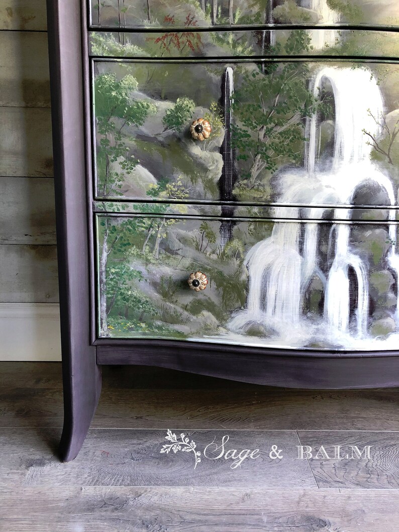 May include: A dark purple dresser with a hand-painted waterfall scene on the drawers. The dresser has a curved bottom and decorative knobs.