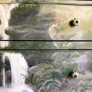 May include: A hand-painted dresser with a waterfall and forest scene. The dresser has three drawers with ceramic knobs. The top drawer has the text "Sage & BALM" on the right side.