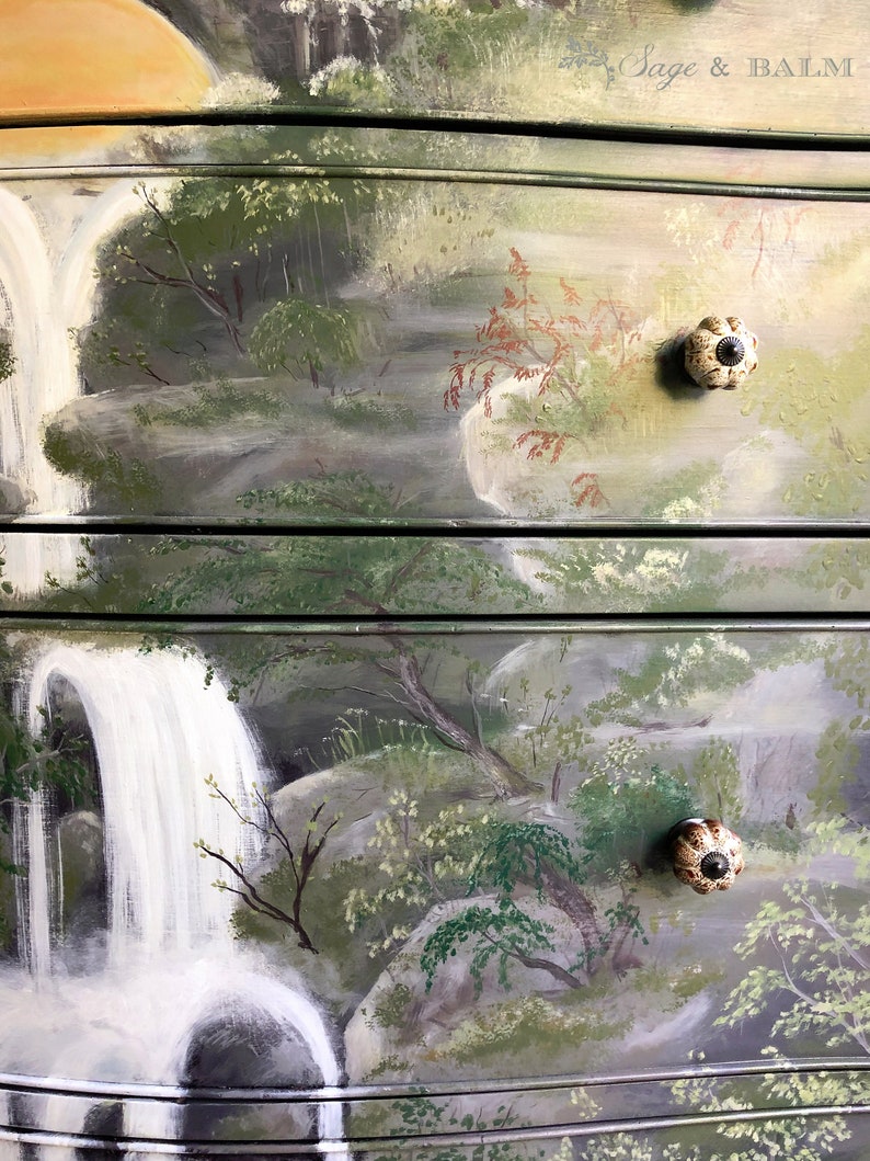 May include: A hand-painted dresser with a waterfall and forest scene. The dresser has three drawers with ceramic knobs. The top drawer has the text "Sage & BALM" on the right side.