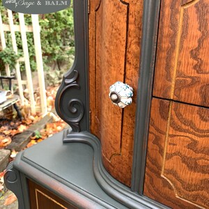 Puede incluir: Un armario de madera vintage con un acabado de pintura verde oscuro. El armario tiene una parte superior curva y detalles ornamentados. Las puertas y los cajones tienen herrajes decorativos con pomos de porcelana blanca.