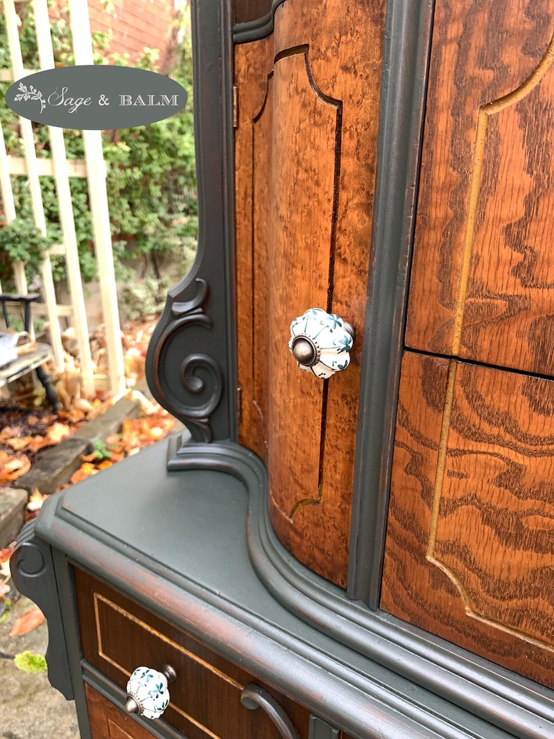 Puede incluir: Un armario de madera vintage con un acabado de pintura verde oscuro. El armario tiene una parte superior curva y detalles ornamentados. Las puertas y los cajones tienen herrajes decorativos con pomos de porcelana blanca.