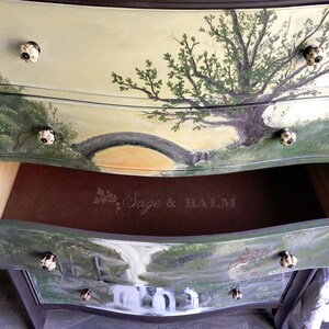 May include: A hand-painted dresser with three drawers features a nature scene on each drawer. The top drawer has a tree with a bridge in the background. The middle drawer is open and shows the brown interior. The bottom drawer has a waterfall scene. Each drawer has a unique knob.